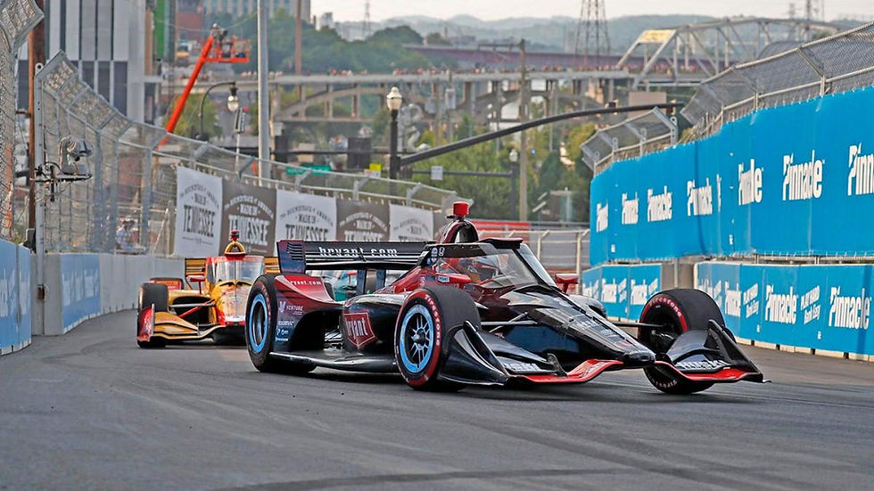 Race cars speeding around a track in a city setting, with advertisements on barriers. The leading car is black and red, followed by a yellow car.