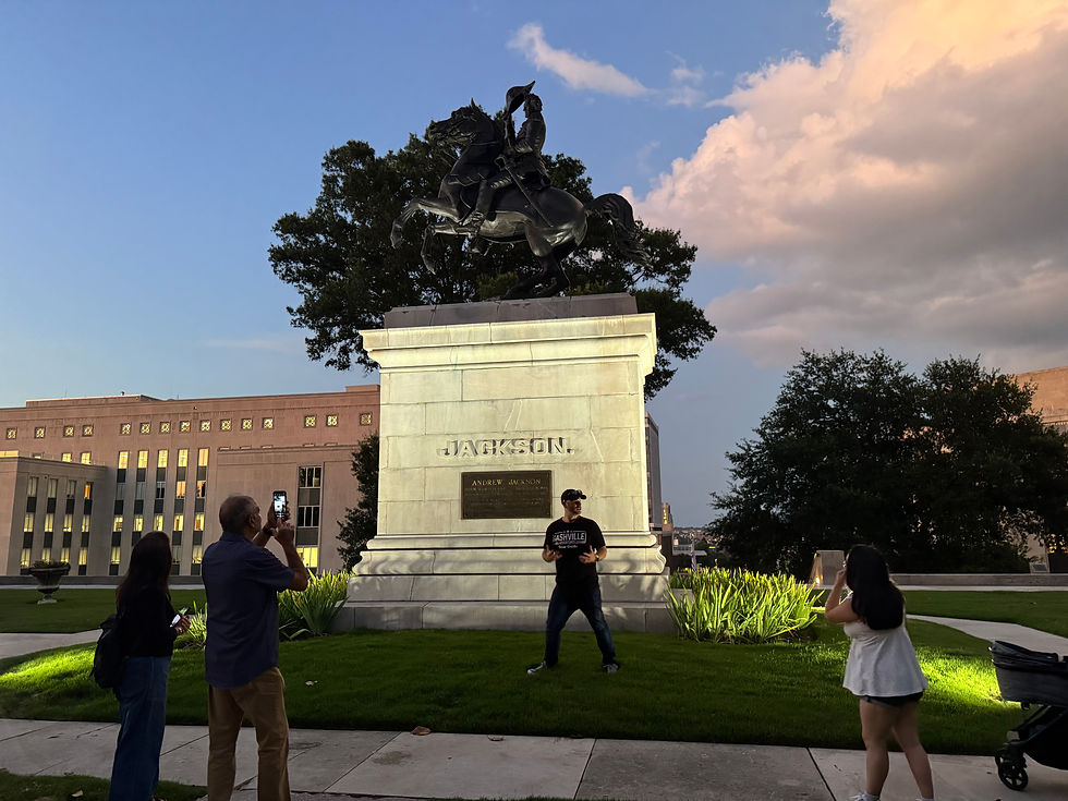 People photographing a man posing near the Andrew Jackson statue in a park. Blue sky, setting sun, and buildings in the background.