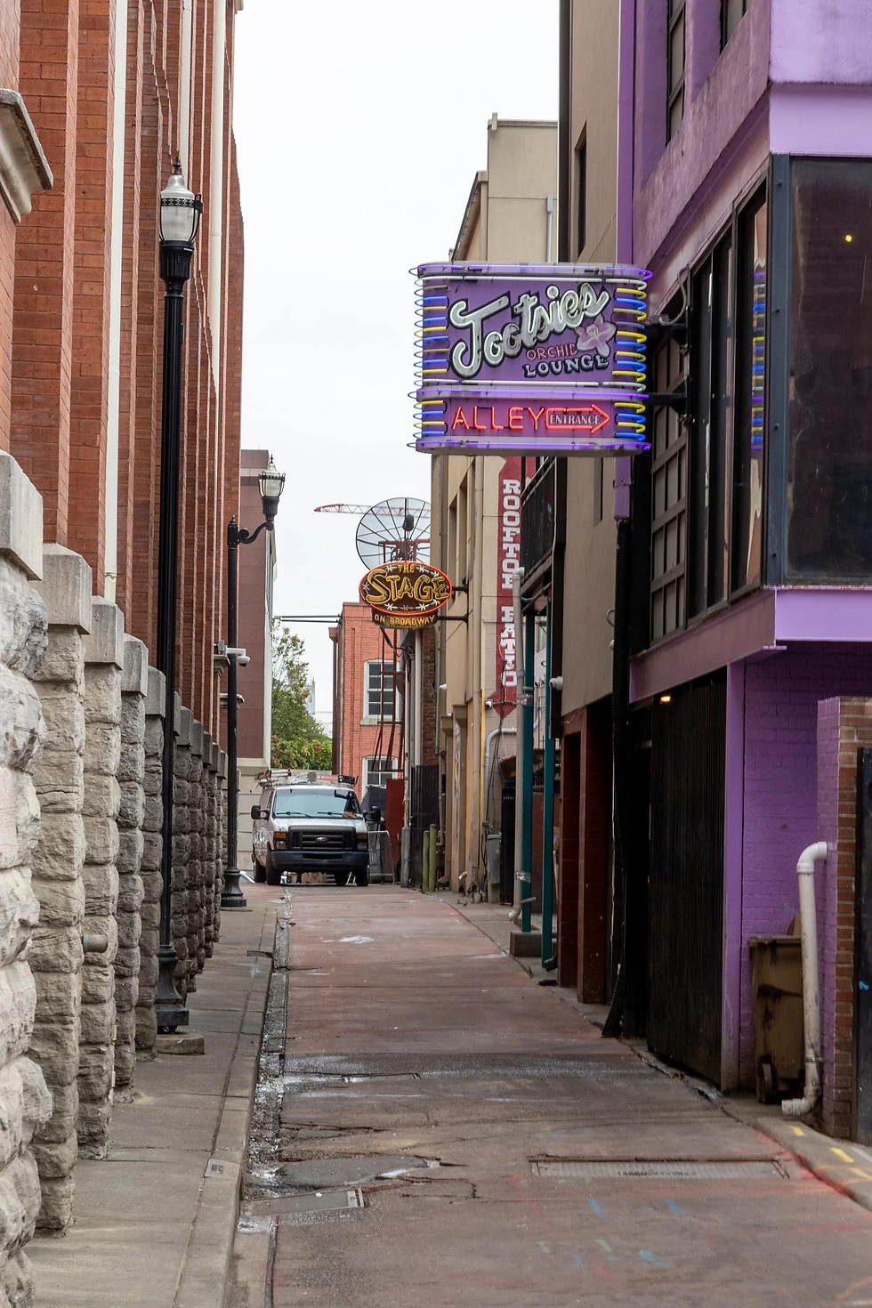 Narrow alley with neon signs for Tootsie's Orchid Lounge. Brick buildings on sides. Parked van in distance. Overcast mood.