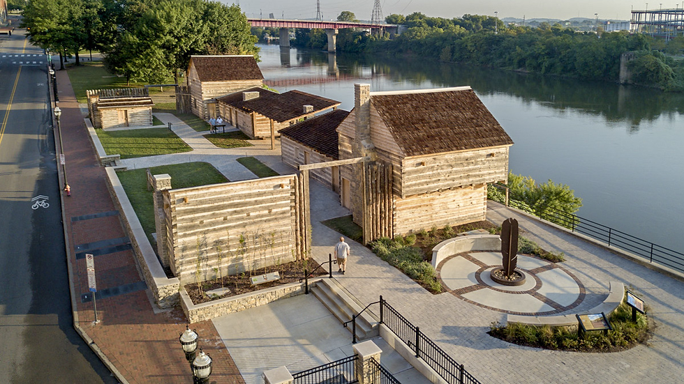 Aerial view of a reconstructed historic fort by a river, featuring wooden buildings, a circular monument, and a person walking on a path.