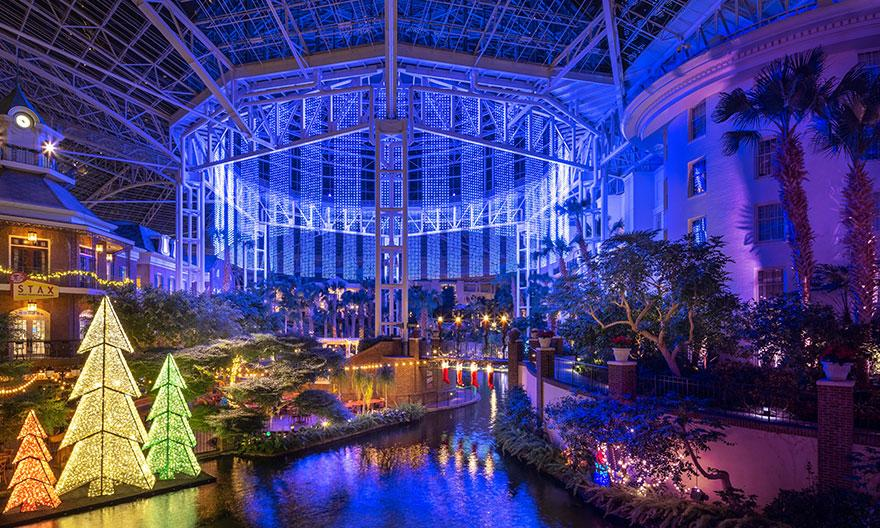 Indoor garden with colorful lit trees by a waterway, under a large glass dome. Blue lights illuminate the structure, creating a festive mood.