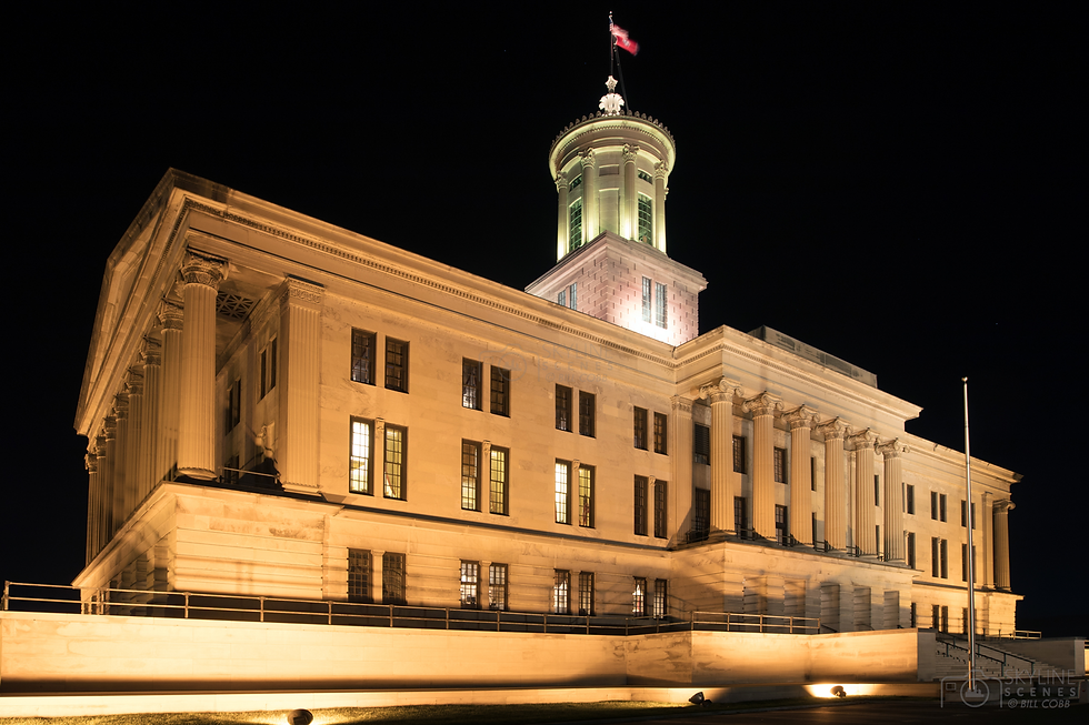 Illuminated neoclassical building with columns and cupola at night. Warm lighting contrasts with the dark sky. Flag on top.