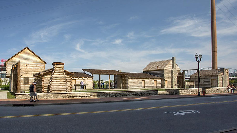 Reconstructed wooden fort with people walking nearby. Clear sky, street in foreground, lamp posts, and bridge visible in the background.