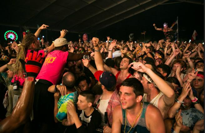 A lively concert crowd with hands raised in excitement. A person in a pink shirt crowd-surfs. Colorful lights are visible in the background.