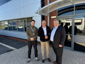 Three men stand outside a large building smiling. Two of them are shaking hands. Glass windows and a brick wall are in the background.