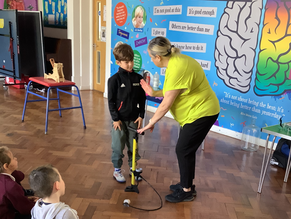 Woman helps boy use a yellow air pump in a colourful classroom with brain-themed wall art. Children watch attentively, sitting cross-legged on the floor.