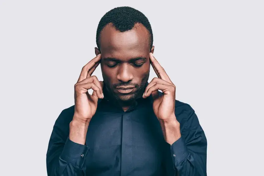 Man in a dark blue shirt touches his temples with fingers, eyes closed, focused expression. Light gray background.