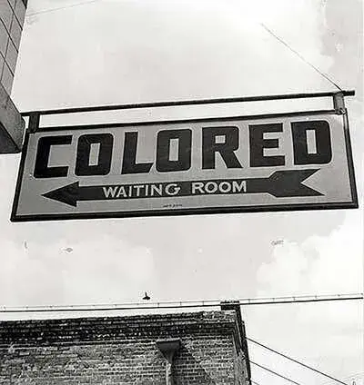 Sign reading "COLORED WAITING ROOM" with arrows, set against a cloudy sky and brick building, highlighting segregation history.