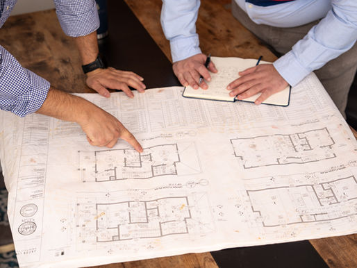 Two team members review architectural plans spread across a wooden table, pointing at floor plans and taking notes.