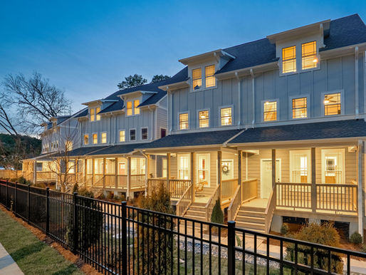 Twilight exterior photo of a row of modern three-story townhomes with gray siding, white trim, and warmly lit windows. Each home features covered front porches and staircases, with a black metal fence running alongside a sidewalk in the foreground. Bare winter trees frame the homes against a deep blue evening sky.