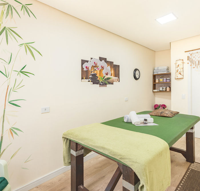 Vista da acolhedora sala de massagens na Pousada Kaliman, na Vila Capivari, com cama de massagens verde, toalhas, um quadro floral na parede e, ao lado, um armário repleto de óleos e cremes essenciais