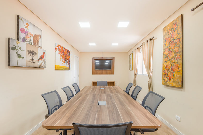 Vista da sala de reuniões com uma grande mesa retangular e ao redor 8 cadeiras, na parede três quadros com imagens de natureza e ao lado painel com televisão embutida na Pousada Kaliman no centro de Campos do Jordão