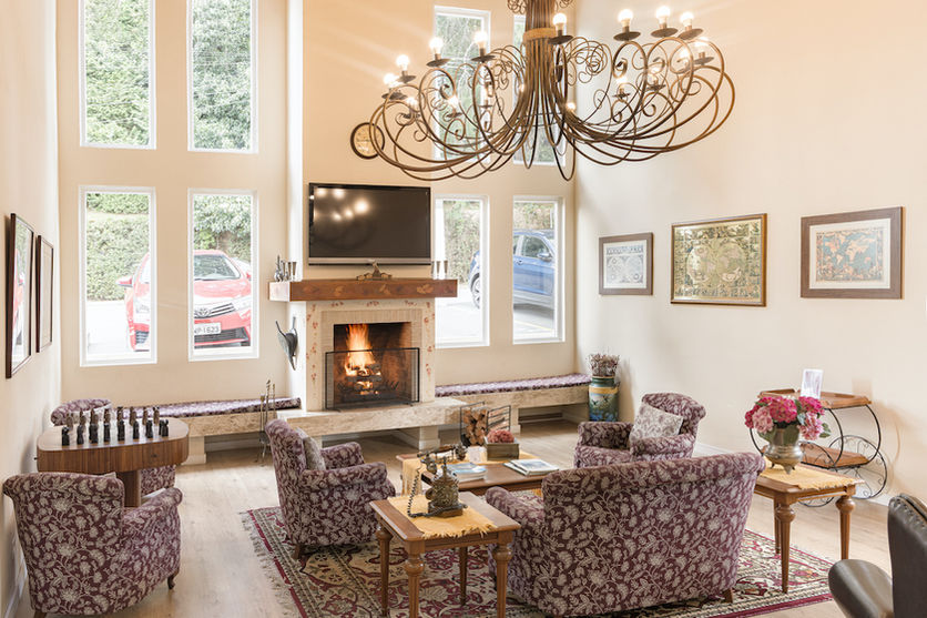 Sala da lareira aconchegante com sofá e poltronas, televisão embutida na parede e abaixo lareira acesso, além de parede com janelas para que os hóspedes possam aproveitar a vista durante a hospedagem em Campos do Jordão