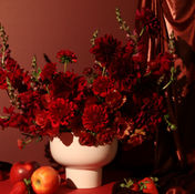 Deep burgundy arrangement with roses, mums, and dark foliage in a white pedestal vase with fruit styling on a dramatic crimson backdrop