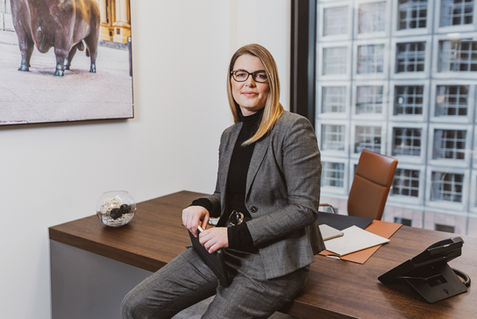 Frau im grauen Anzug sitzt entspannt am Schreibtisch vor einer modernen Fensterfassade – stilvolles Businessporträt in Büroatmosphäre.