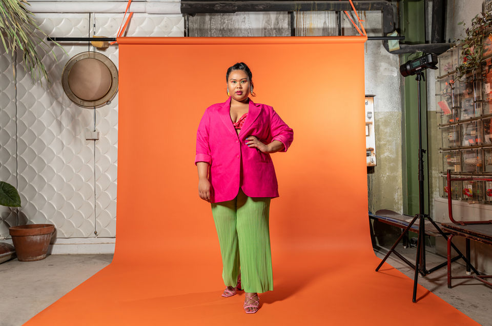 Selbstbewusste Frau in pinkem Blazer und grüner Hose posiert vor orangefarbenem Fotohintergrund in einem Studio.