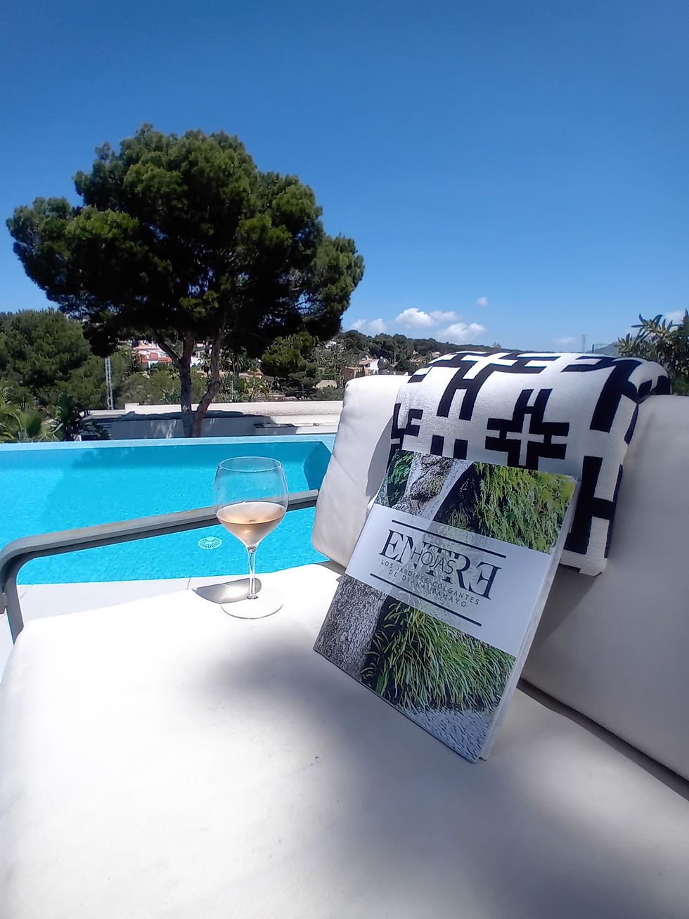 Fotografía cenital y cálida del libro EntreHojas sobre una silla al lado de una piscina y una copa de vino