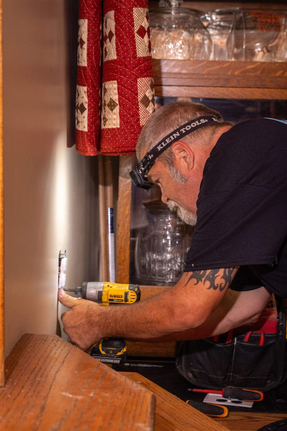 Man with Klein Tools headlamp uses a DeWalt drill on a wall in a cozy kitchen, with a red quilt hanging nearby.