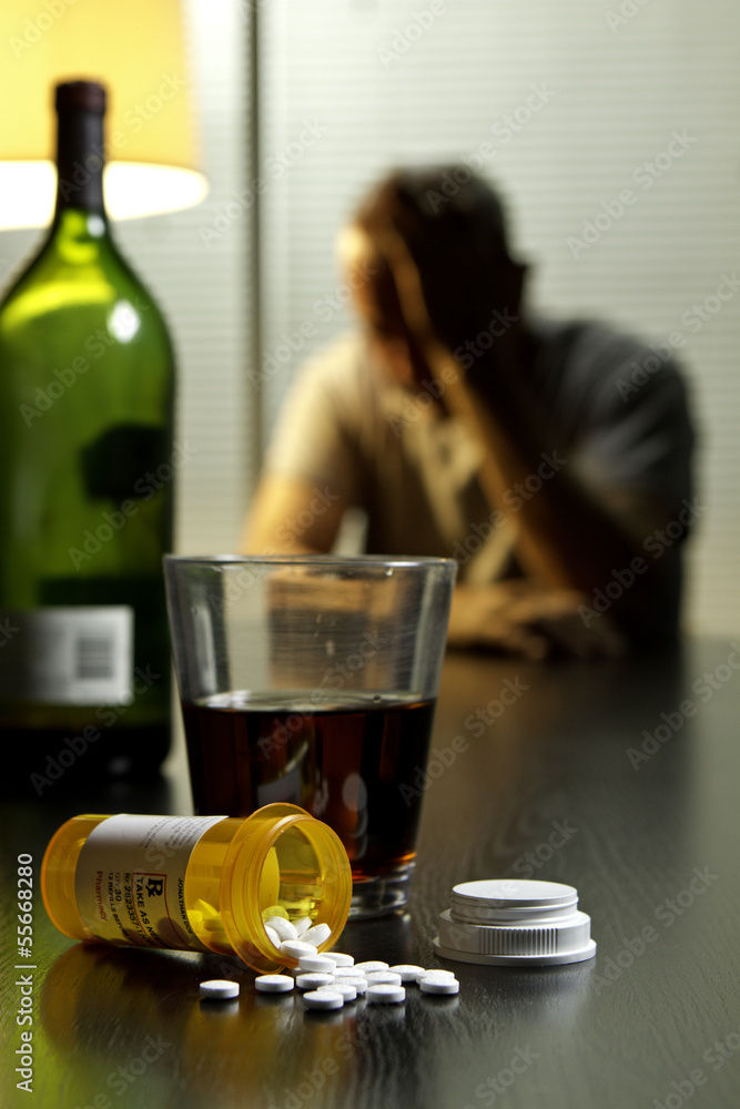 A man with his head in hands in front of a bottle of wine and drugs.