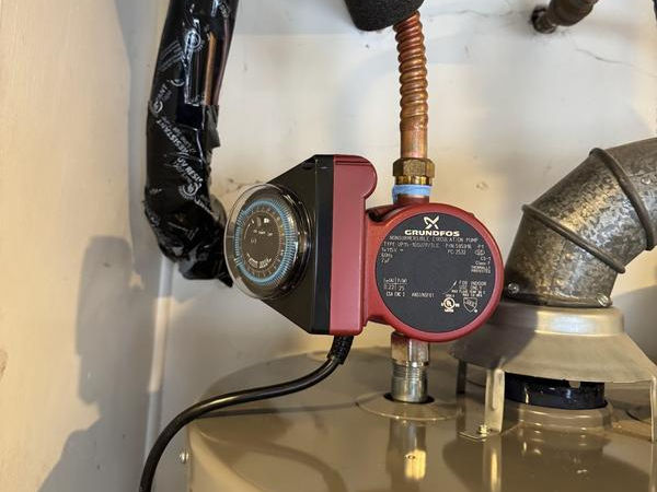 Grundfos circulation pump with timer attached to a water heater, surrounded by pipes and ducting in a utility closet.