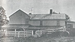 A black-and-white photo of an old farmhouse with a tin roof, surrounded by a wooden fence. Trees in the background create a rural setting.