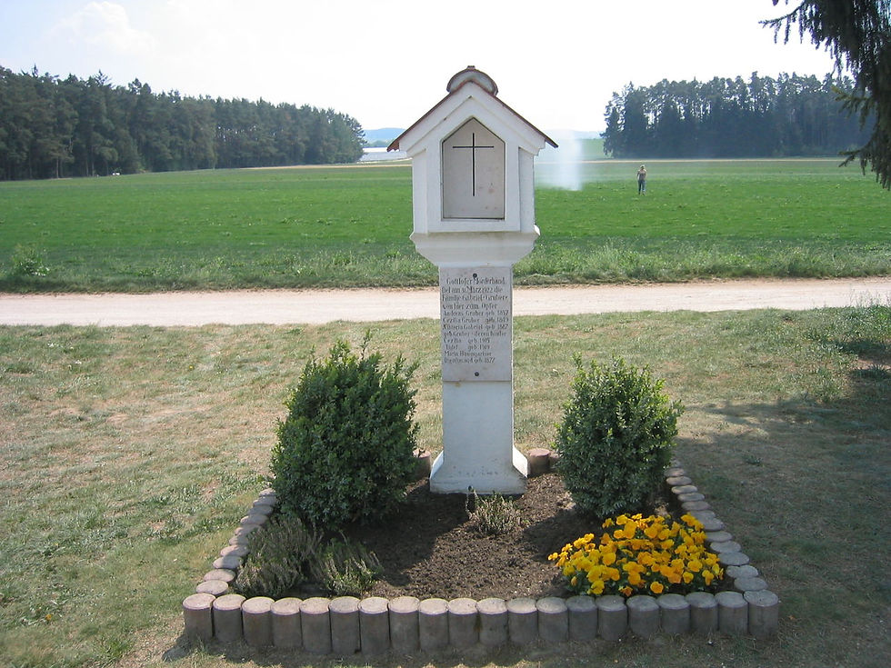 White shrine with a cross stands in a grassy field, surrounded by bushes and yellow flowers. A dirt path and trees are in the background.