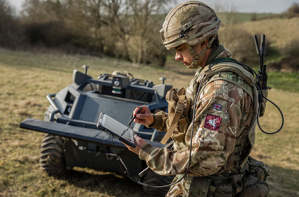 Soldier in camouflage uses a tablet beside an UGV in a grassy field, focused on a military task.