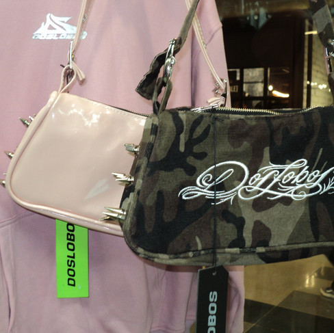 Dos bolsos exhibidos en una tienda - uno es de color rosa pálido/nude con tachuelas metálicas puntiagudas, y el otro tiene un patrón de camuflaje con "Dobobs" bordado. Ambos tienen etiquetas de precio verdes "DOBOBS" y están exhibidos contra un fondo rosa.