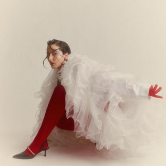 Fotografía artística de una persona en pose dinámica contra fondo beige claro, vistiendo un conjunto rojo con zapatos de tacón, y una extravagante pieza de tul blanco que envuelve parcialmente su cuerpo como una capa voluminosa