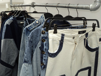 Un perchero con prendas de mezclilla y pantalones blancos. El perchero muestra varias chaquetas vaqueras en diferentes tonos y un par de pantalones blancos con acentos de mezclilla azul. Los pantalones blancos tienen una etiqueta "KATTO JEANS" y parecen ser talla 40.