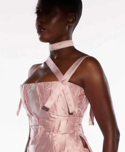Un primer plano de una persona con pelo muy corto, vistiendo un top rosa claro con tirantes y un collar a juego del mismo color. La prenda tiene un diseño estructurado con detalles en el escote. La imagen está tomada contra un fondo blanco, mostrando solo la parte superior del torso y el perfil del rostro.
