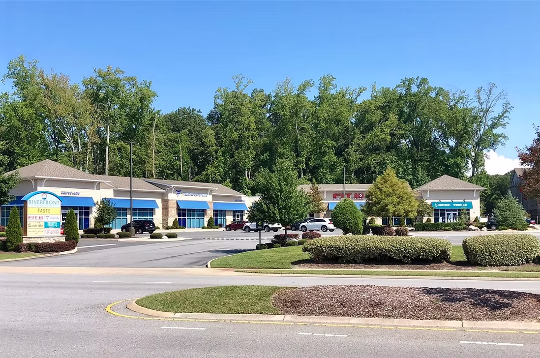 Riverfront Shoppes street view with retail buildings, tenant signage, landscaped medians, parking, and wooded backdrop