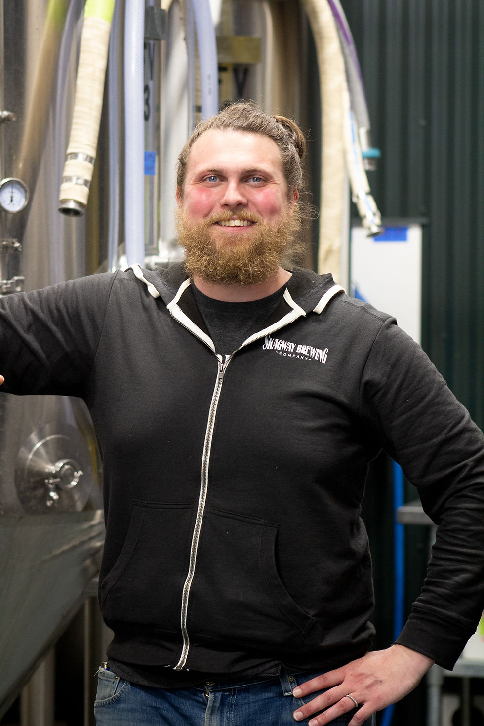 A tall man with a robust beard and a cheerful expression looks into the camera, standing next to a stainless steel tank which is draped with heavy-duty hoses.