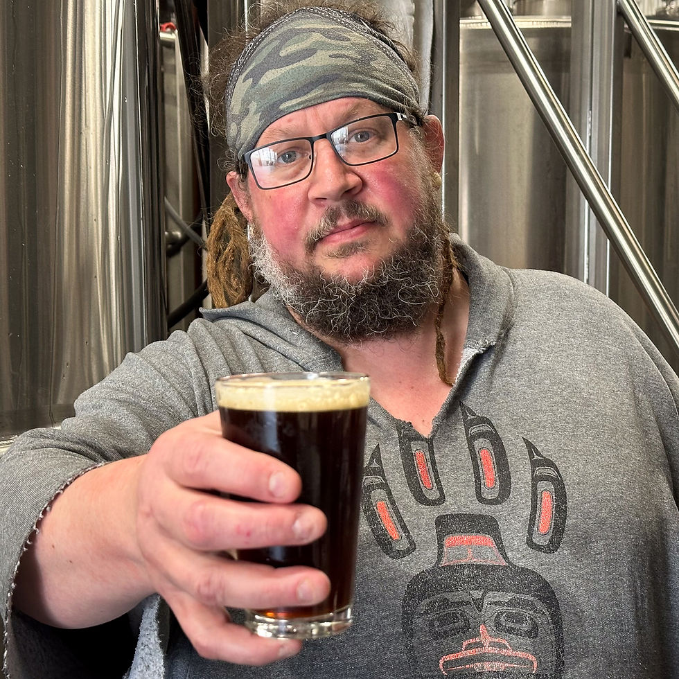 A bearded man with glasses, a bandana, and a sweatshirt featuring a formline-style paw stands in front of stainless steel tanks, holding up a glass of dark beer