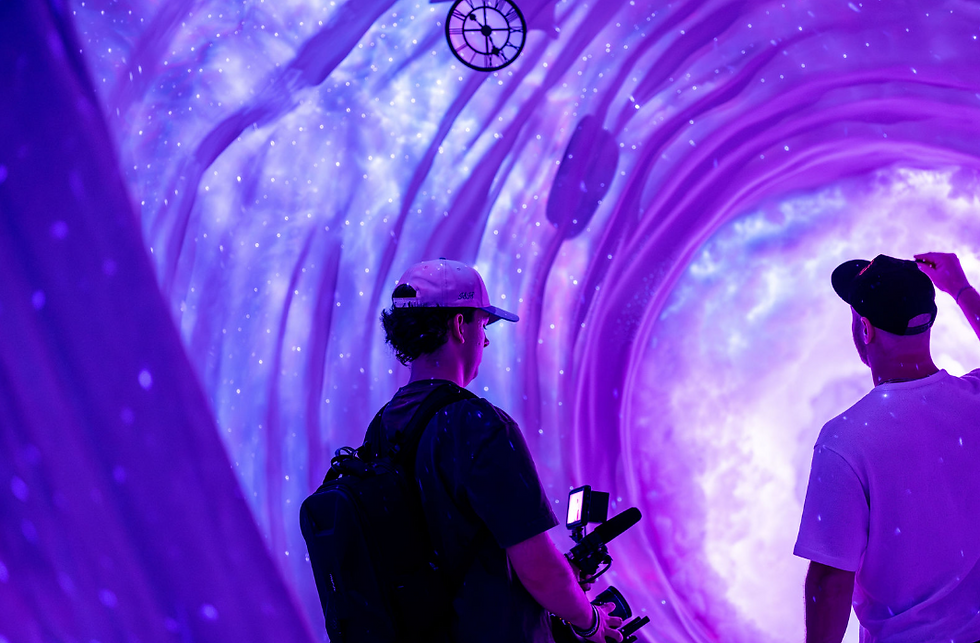 Two men view a glowing purple tunnel-like light installation, one holding a camera rig, with a starry swirl backdrop.