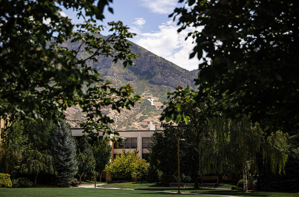 Tree-framed campus lawn with a building, mountain hillside marked by a white Y, under a bright blue sky.