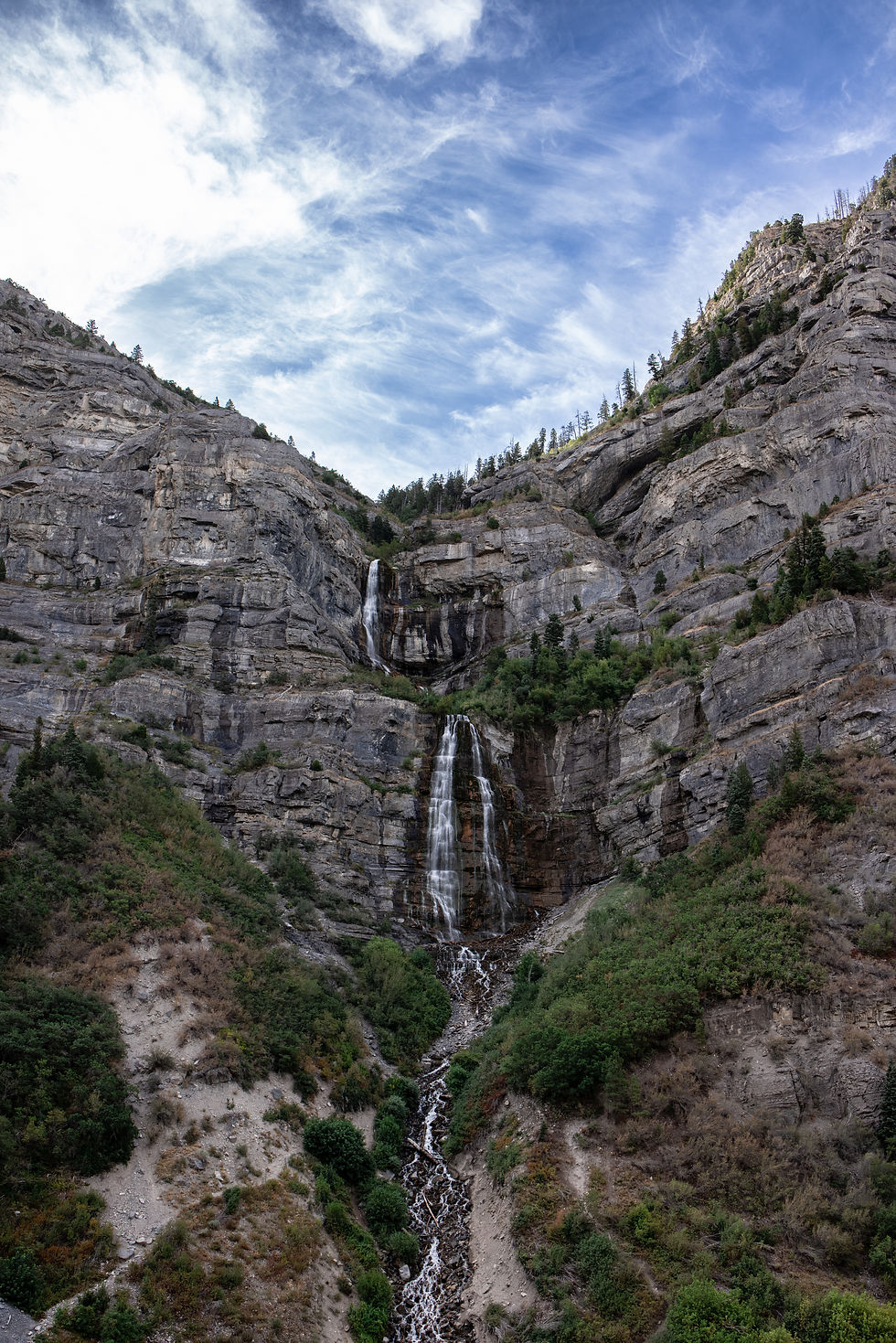 Tall waterfall cascading down rocky canyon cliffs under a blue, wispy-cloud sky, with green shrubs on steep slopes.