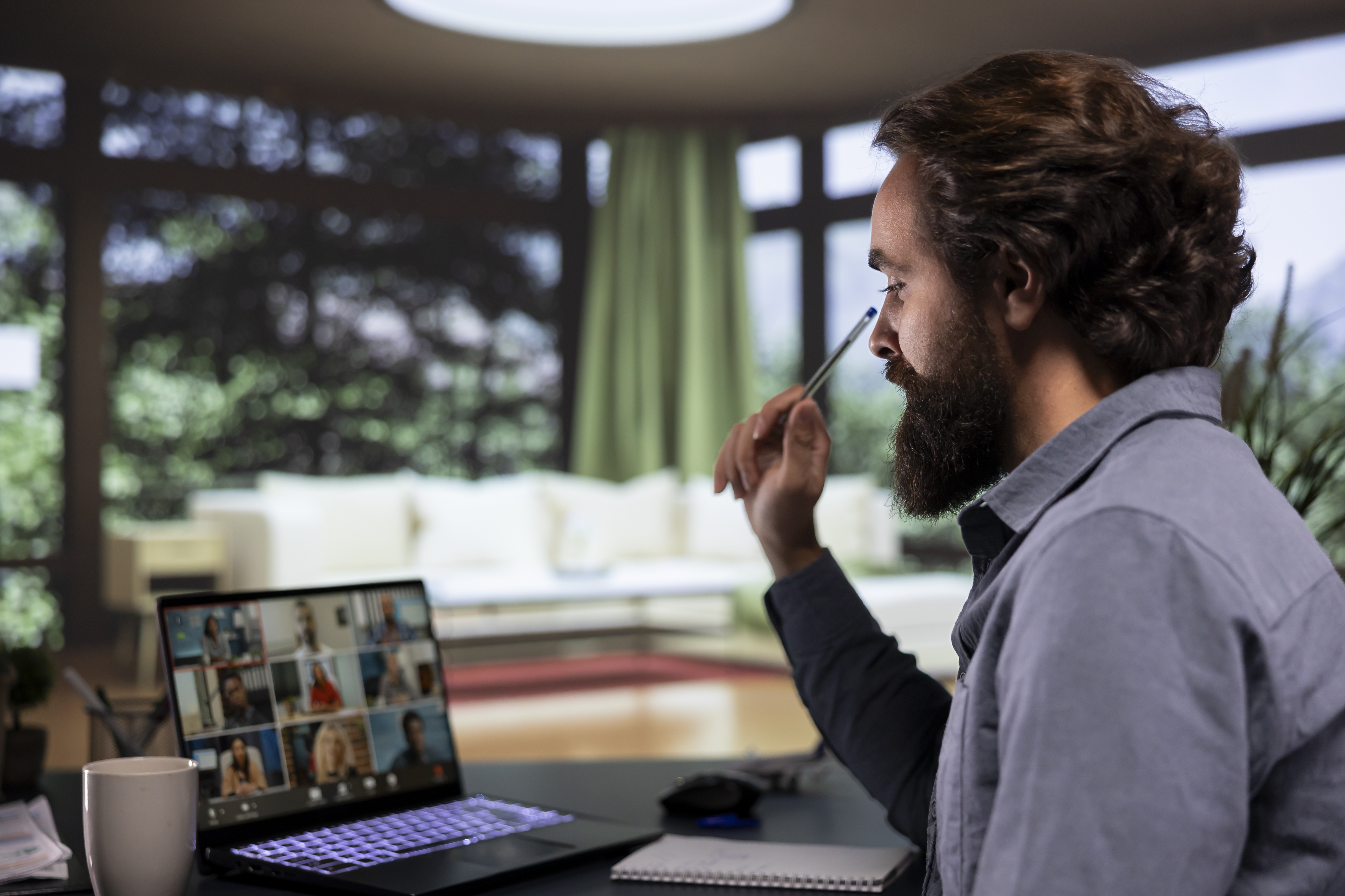 Man with a beard in a blue shirt attends a video call on a laptop. He's holding a pen in a bright, homey setting with a mug nearby.