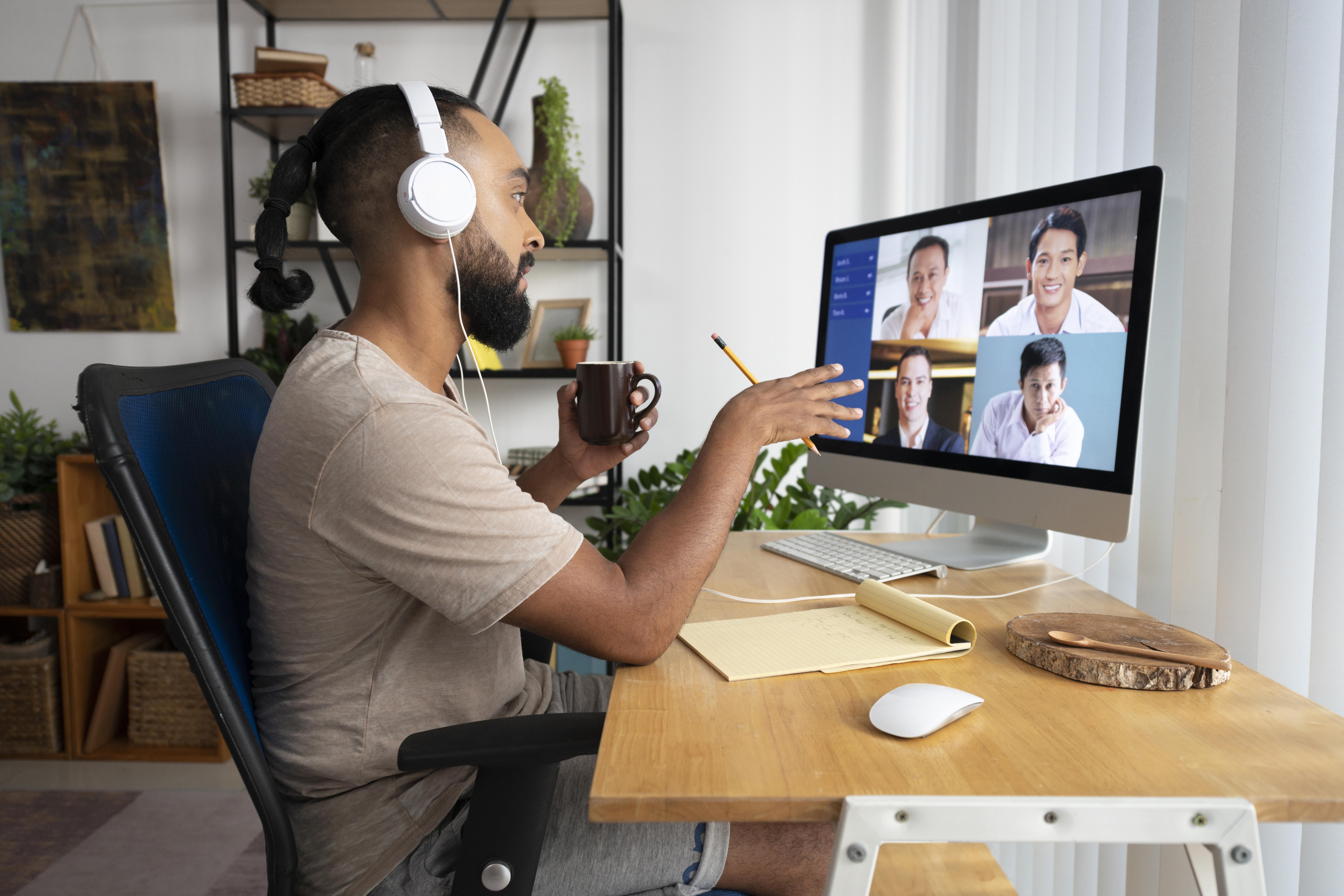 Man with headphones holds a mug, video conferencing on a desktop. Notepad and pencil on wooden desk. Bookshelf and plants in background.
