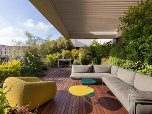 מרפסת גג מעוצבת עם דק עץ, ספה אפורה מודרנית, כורסה צהובה ושולחנות קפה צבעוניים, מוקפת בצמחייה ירוקה ושופעת תחת פרגולה.