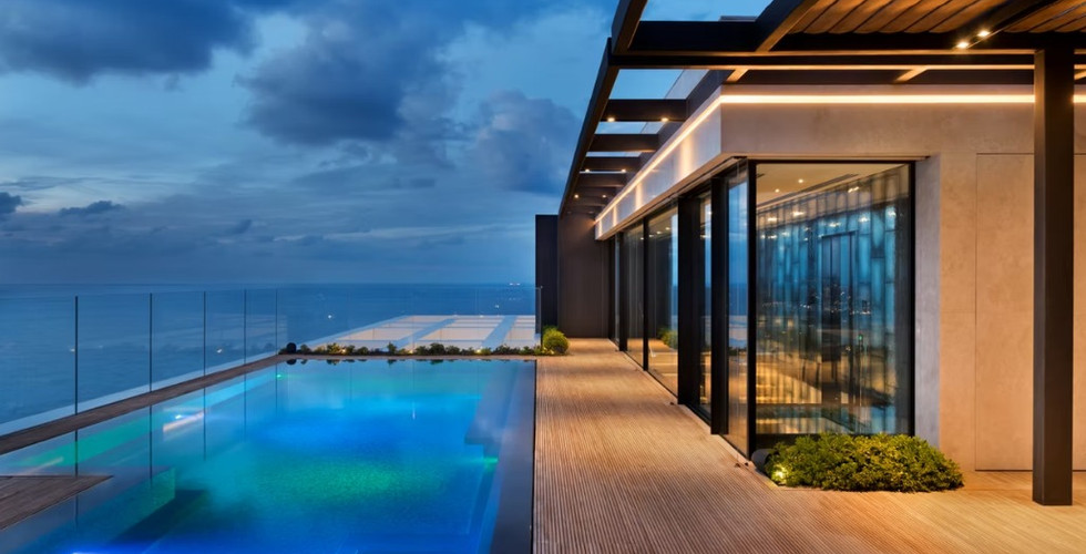 פנטהאוס יוקרתי עם בריכת אינפיניטי מוארת, דק עץ ונוף פתוח לים בשעת ערב, עם תאורה אדריכלית מודרנית וצמחייה משולבת