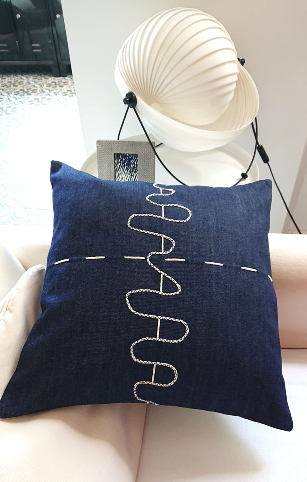 CELESTO Housse de coussin Déco jean brut, présentée sur un canapé écru, à côté d'une jolie lampe.