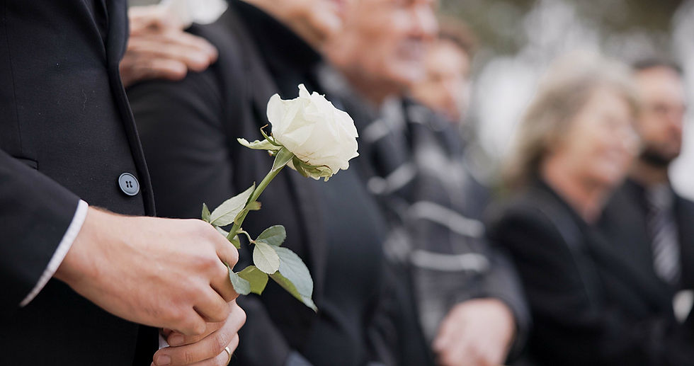 Close up of hands holding a white flower with people in the background wearing black in a funeral.