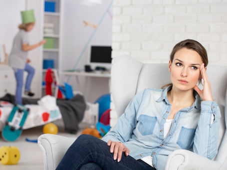 Worried woman sits in a white armchair in a messy child’s room, while a child plays in the background.