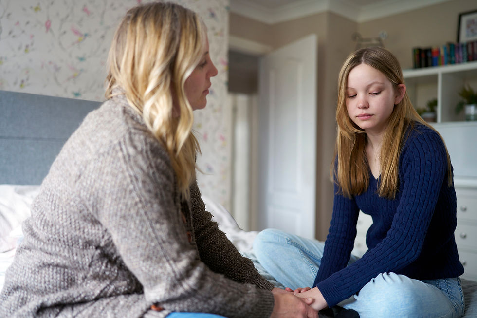 A mother and her teenage daughter sit on a bed holding hands, one comforting the other in a softly lit bedroom, both looking solemn.