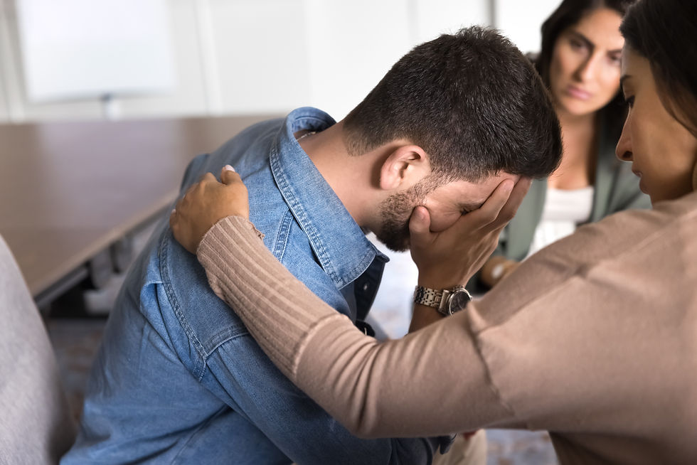 Grieving man with his face buried in his hands and women consoling him with her hand on his shoulder during group therapy session.