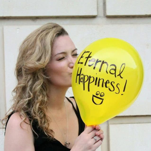 Woman kisses a yellow balloon with "Eternal Happiness!" text and a smiley face. Background is a light-colored wall. Mood is joyful.