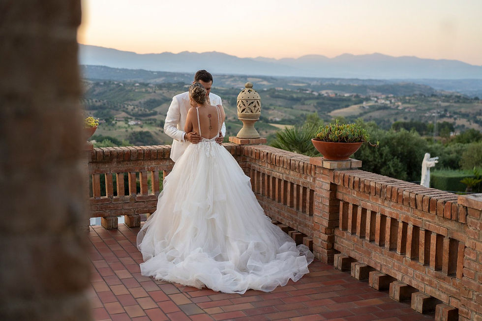 Bride and groom embrace on a brick terrace overlooking scenic hills at sunset. Bride wears a flowing white gown, creating a romantic mood.