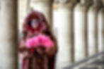 Person in elaborate red and pink costume and mask, holding a pink feather fan. Stone columns in background, creating an elegant, mysterious mood.