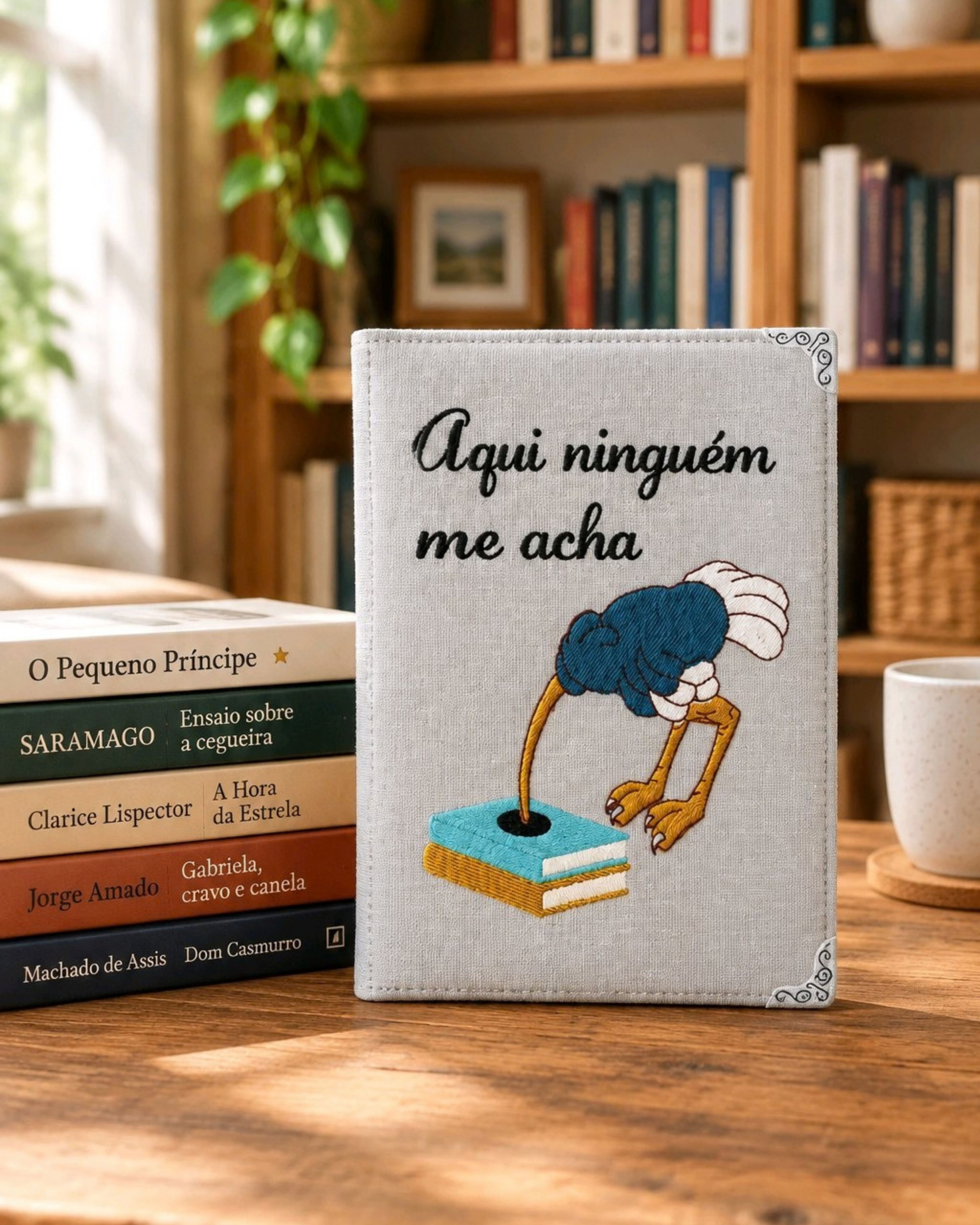 Capa Kindle Aqui Ninguém Me Acha Cinza Nevoeiro, com frase bordada e avestruz de cabeça enfiada em pilha de livros
